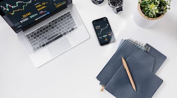 Aerial view of a laptop and smartphone displaying finance data, with notebooks, pen, and a plant on a desk.