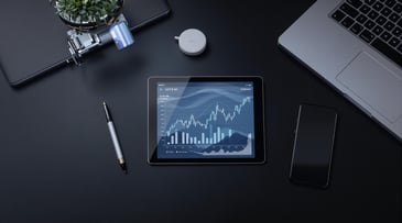 Top view of a tablet displaying stock market graphs on a sleek workspace with a laptop, smartphone, and pen.