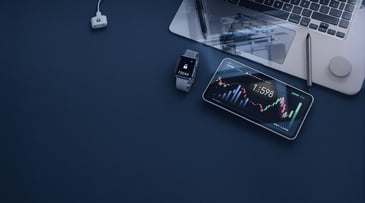 A workspace scene featuring a smartphone displaying stock market data, a smart watch, laptop, and pen on a blue surface.