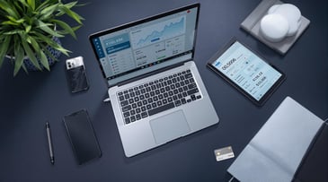 Aerial view of a laptop and tablet displaying financial dashboards, surrounded by a phone, pen, and plant.
